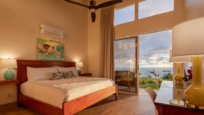 A bedroom with a wooden bed, a painting, an air conditioner, a balcony with a view of the ocean, and a dining area with a table and chairs. - Luxury retreat with panoramic ocean view, private infinity pool, concierge (Uvita)