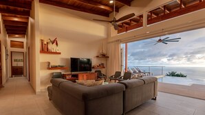 A spacious living room with a large gray sofa, a television, and a dining area with chairs. There is a ceiling fan and a view of the ocean through large windows.