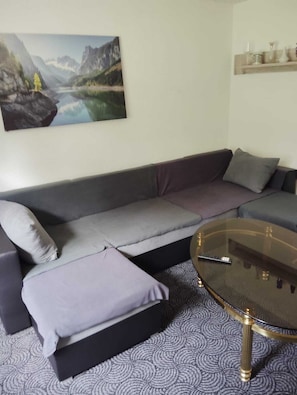 A modern living room with a grey sofa, a glass coffee table, and a painting of a mountain landscape on the wall.