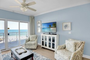Relax in this inviting living room with a stunning beach view through large glass doors, featuring a ceiling fan, flat-screen TV, and comfortable armchairs.