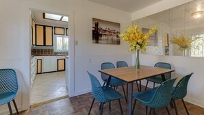 Een eethoek met een houten tafel en blauwe stoelen, een vaas met gele bloemen, en een keuken op de achtergrond.