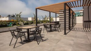 An outdoor patio with tables and chairs, a wooden pergola, and a view of the cityscape.