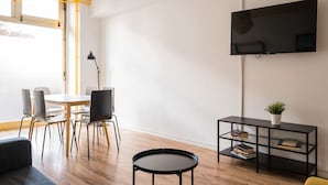 A modern living room with a dining area, a flat-screen TV mounted on the wall, a black bookshelf, and a round coffee table.