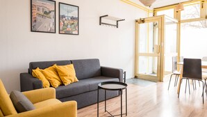 A modern living room with a grey sofa, a yellow armchair, and a small round table. There are two framed cityscape posters on the wall, a hanging light fixture, and a door leading to another room.