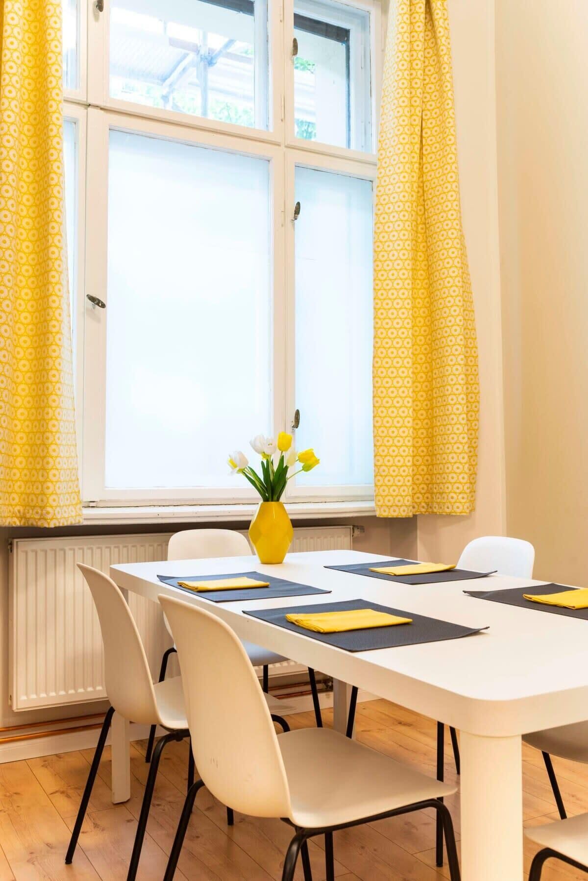 A dining area with a white table, yellow patterned curtains, and a vase with flowers.