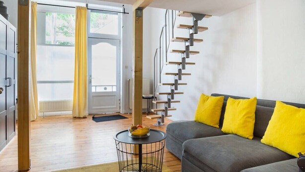 Un salon moderne avec un canapé gris, des coussins jaunes, une table ronde noire et un escalier en colimaçon.