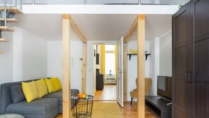 Un appartement moderne avec un canapé gris, des coussins jaunes, une petite table avec des fruits, un téléviseur et un escalier en bois.