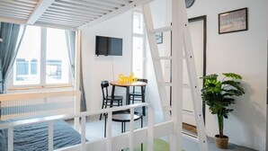 Een studentenkamer met een stapelbed, een bureau met een stoel, een kleine tafel met een bordje 'Smile', een bloempot en een televisie aan de muur.