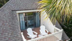 A deck with two white Adirondack chairs and a small table, overlooking a covered patio area with a glass door.