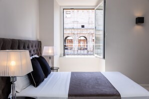 A hotel room with a bed, a lamp, a nightstand, and a window with a view of a building.