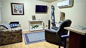 Un salon comprenant un canapé, un fauteuil et une petite table avec une lampe. Un téléviseur est fixé au mur, et un tableau encadré est accroché au-dessus.