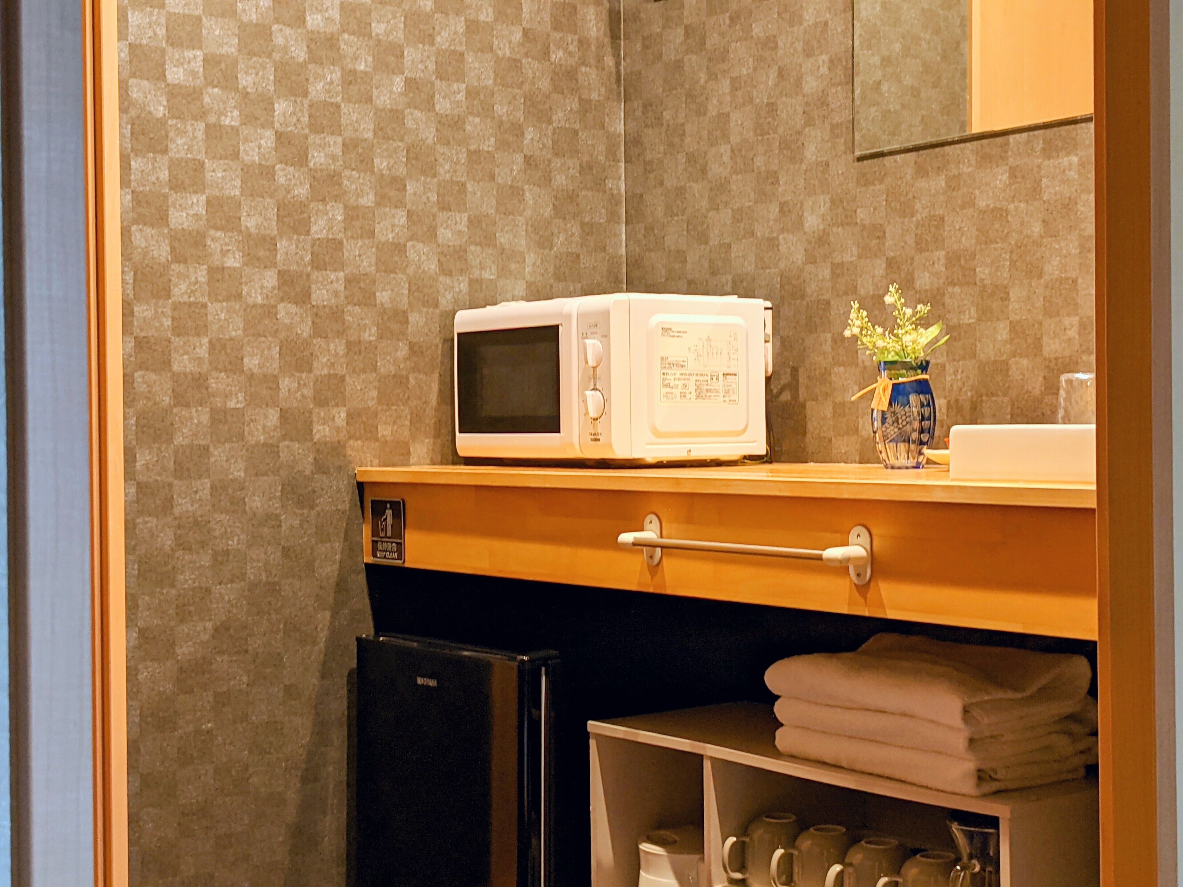 A kitchenette with a microwave on the counter, a small refrigerator, and a shelf with towels and kitchenware.
