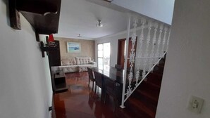 A spacious living room with a wooden floor, a sofa, and a dining area with a table and chairs. - Cobertura Duplex - Santos (Santos)