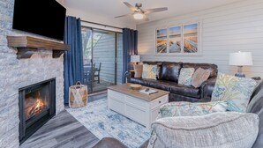 A living room with a fireplace, a leather sofa, a coffee table, and a large window with curtains. - 18C Richmond Park, remodeled villa with full access to amenities (Myrtle Beach)
