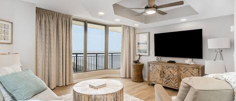 A spacious living room with a large TV, comfortable seating, a ceiling fan, and a view of the outdoors.