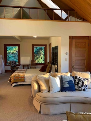 A spacious loft bedroom with a large bed, a sofa, and a view of the outdoors through multiple windows.