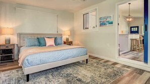 Un dormitorio con cama, lámparas de noche, ventilador de techo, ventana y puerta que da a otra habitación.