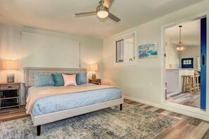 Un dormitorio con cama, lámparas de noche, ventilador de techo, ventana y puerta que da a otra habitación.