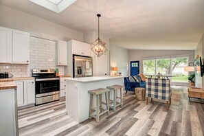 A modern kitchen with white cabinets, a central island, bar stools, and a blue sofa in a bright living area with large windows. - Boji Waterfront Retreat - Millers Bay Canals (Milford)