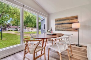 A dining area with a round table, white chairs, and a view of a residential neighborhood through large windows.