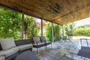 Patio esterno con zona salotto coperta, pavimento in pietra e vegetazione circostante.