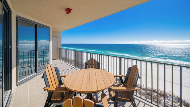 A balcony with a round wooden table and chairs, overlooking a beach with the ocean. - Exquisite 3 Bedroom Condo at Phoenix X 1006 (Orange Beach)