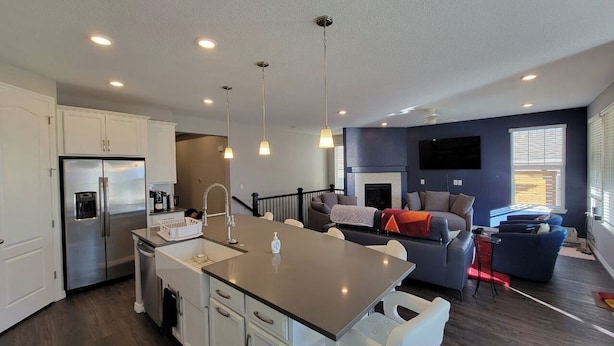 A modern kitchen with a central island, white cabinetry, and stainless steel appliances. There is a living area with a sofa, a fireplace, and a TV mounted on the wall.