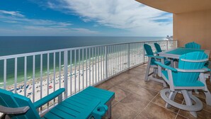 A balcony with turquoise chairs and a table, overlooking a beach with a white railing and a view of the ocean.