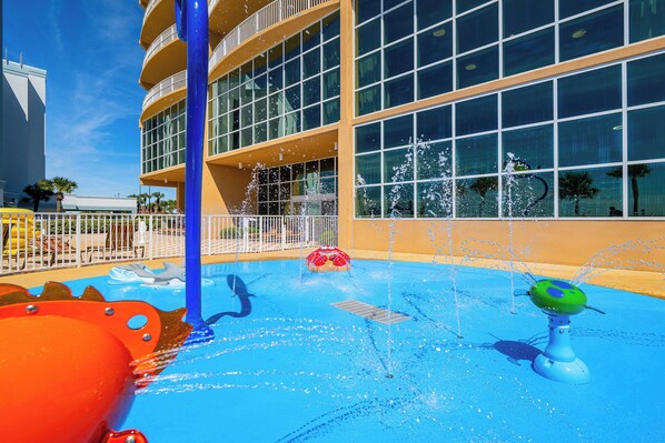 A water play area with a blue pool, a large building with balconies, and a white fence.