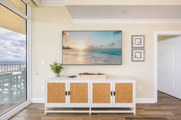 A room with a large window overlooking a beach, a white cabinet with woven doors, a framed painting of a sunset, and a potted plant on the cabinet.