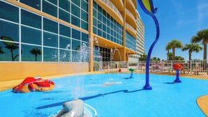Un área de juegos acuáticos con una escultura de delfín, palmeras y un edificio con grandes ventanales.