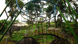 A playground with a circular climbing structure, a bridge, and large rocks. - Fort Munnar (Devikolam)