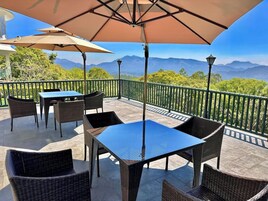 Un patio exterior con mobiliario de mimbre y una mesa, un paraguas grande y vistas a montañas.