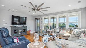 A spacious living room with a ceiling fan, a flat-screen TV, a leather sofa, a patterned armchair, and a large window with a view. - Beach 45: Large, Modern Home Situated on the Davis Canal, Around 500 Feet to Bea (Oak Island)