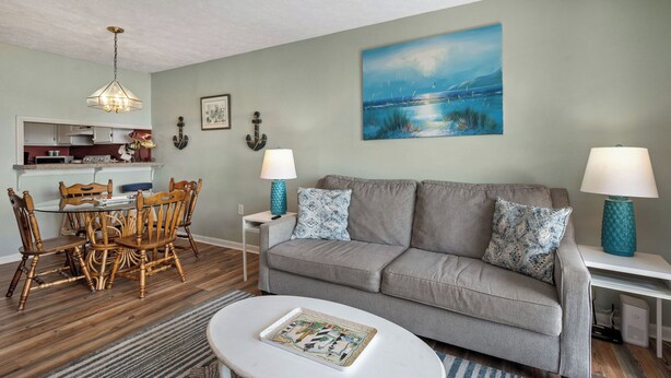 A living room with a grey sofa, two side tables with lamps, a dining table with chairs, a coffee table, and a painting on the wall.