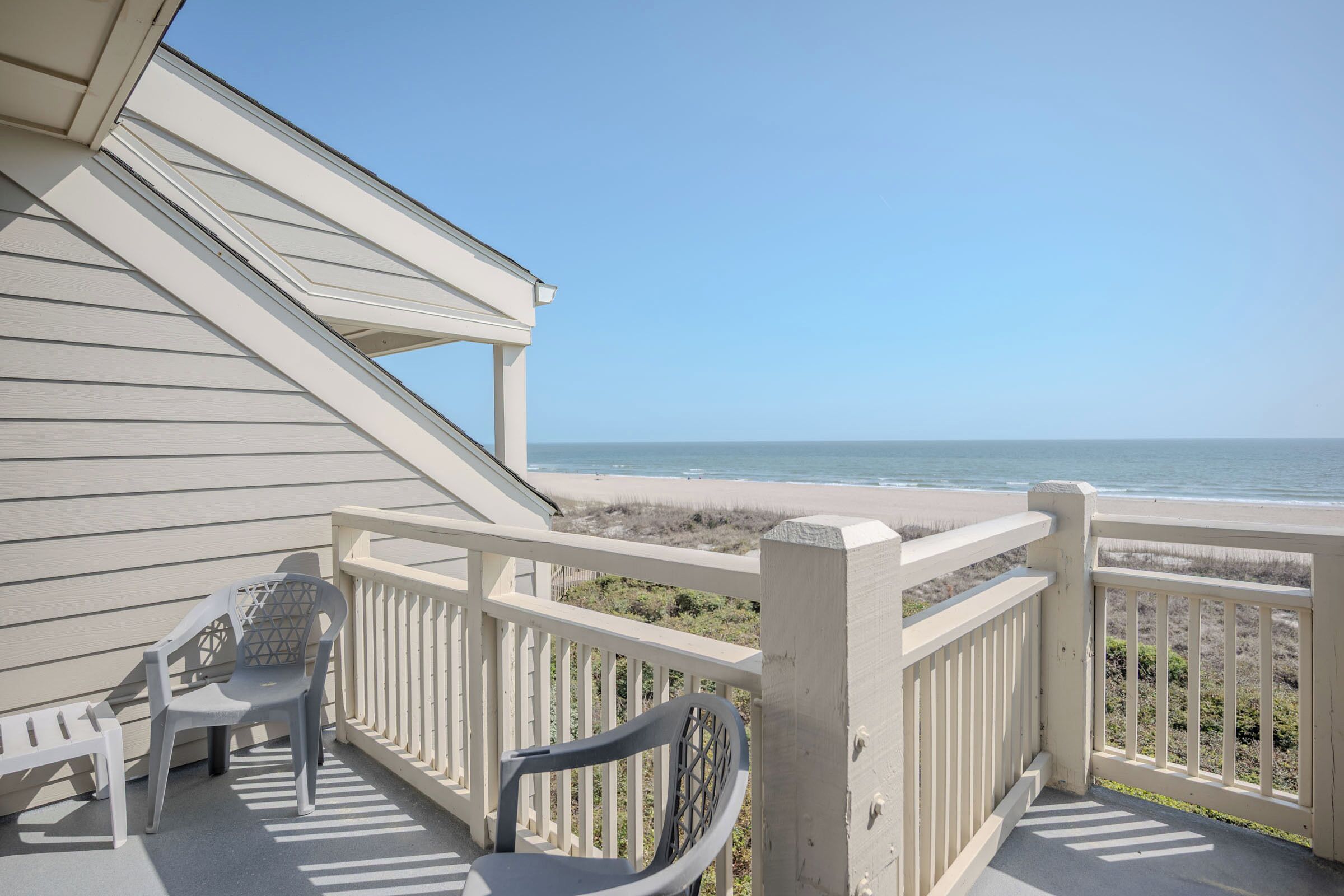 A balcony with a view of the beach, a white railing, and two plastic chairs.