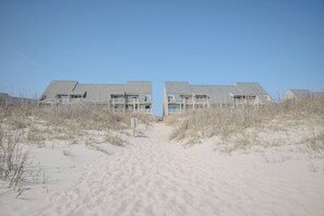 A beachfront hotel with a symmetrical facade, a central entrance, and a sandy path leading to the building. - Anchors Away: Oceanfront Condo with Community Pool, Close to Oak Island Pier (Caswell Beach)