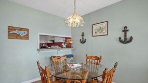 A dining area with a round glass table surrounded by wooden chairs, a kitchen in the background, and nautical-themed wall decor.