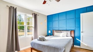 Une chambre avec un grand lit, une fenêtre avec des rideaux, un mur d’accent bleu et un ventilateur de plafond.