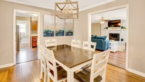 Un coin repas avec une table en bois et des chaises blanches, un lustre et une vue sur la cuisine.