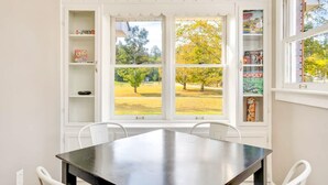 Un coin repas avec une table et des chaises, une fenêtre donnant sur des arbres et un parc, et des étagères intégrées où sont rangés des jeux de société.