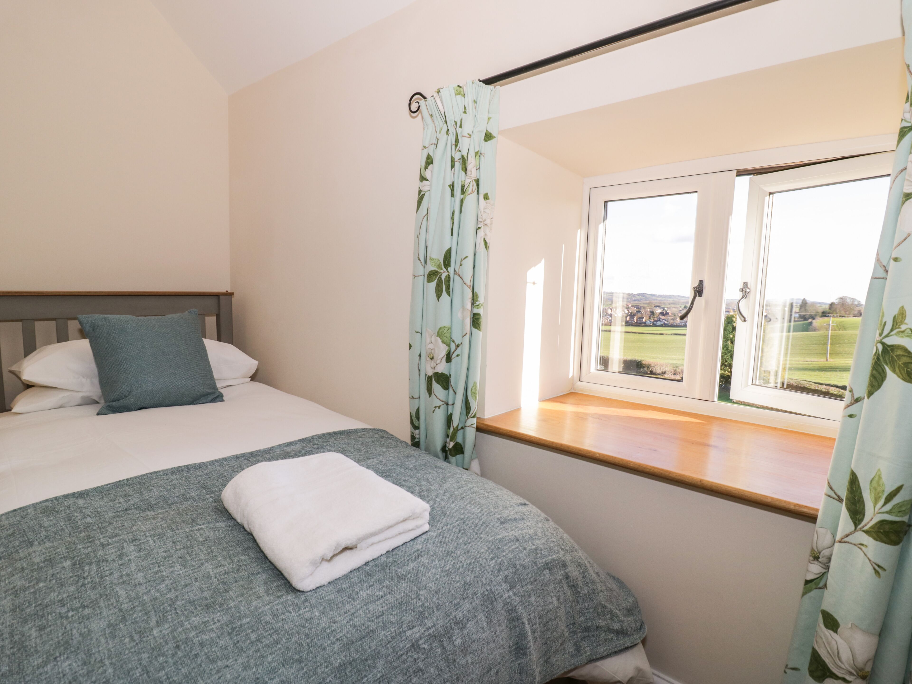 Una cama bien hecha con un edredón gris y una almohada blanca, una ventana con vistas a un paisaje verde y una cortina con estampado floral.