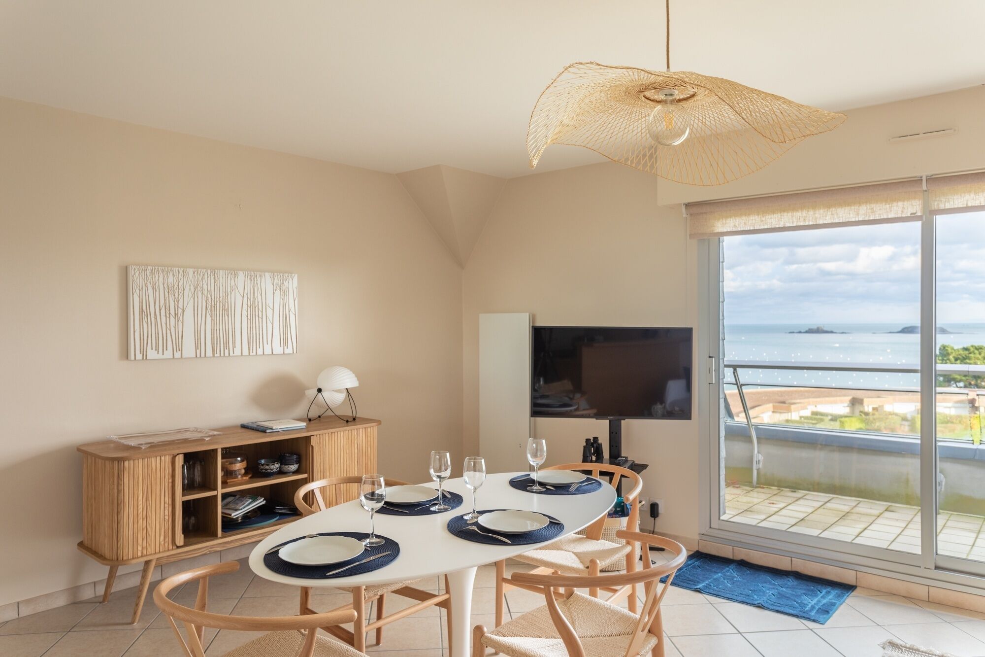 A dining area with a table set for four, a television, and a view of the sea through a sliding glass door.