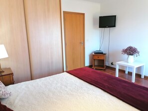Ein Schlafzimmer mit einem Bett, einem Holzschrank, einem an der Wand montierten Fernseher, einem kleinen Tisch mit Blumen in einer Vase und einer Lampe auf einem Nachttisch.