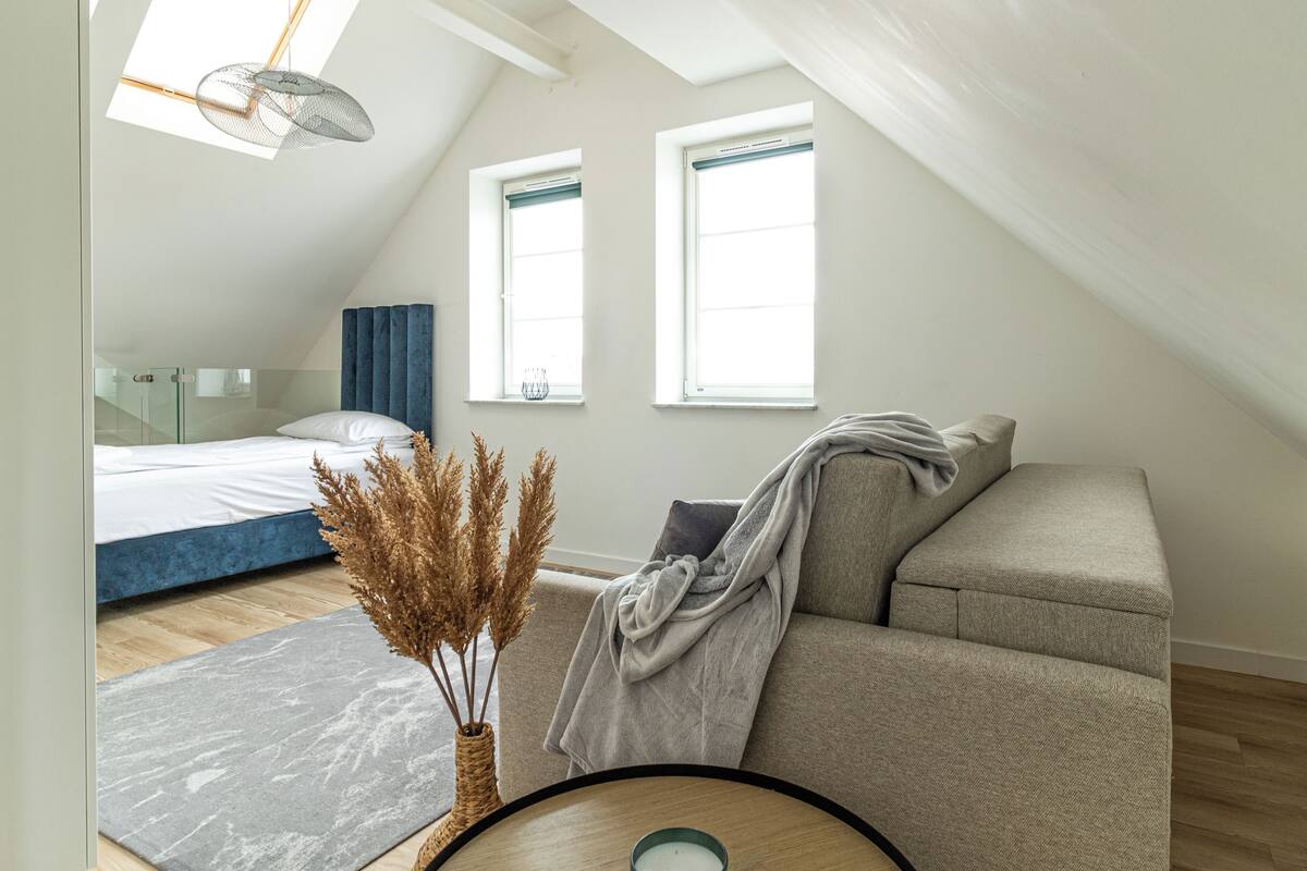 A modern, well-lit room with a bed, a sofa, and a small table with a candle. There are two windows providing natural light and a skylight above the bed.