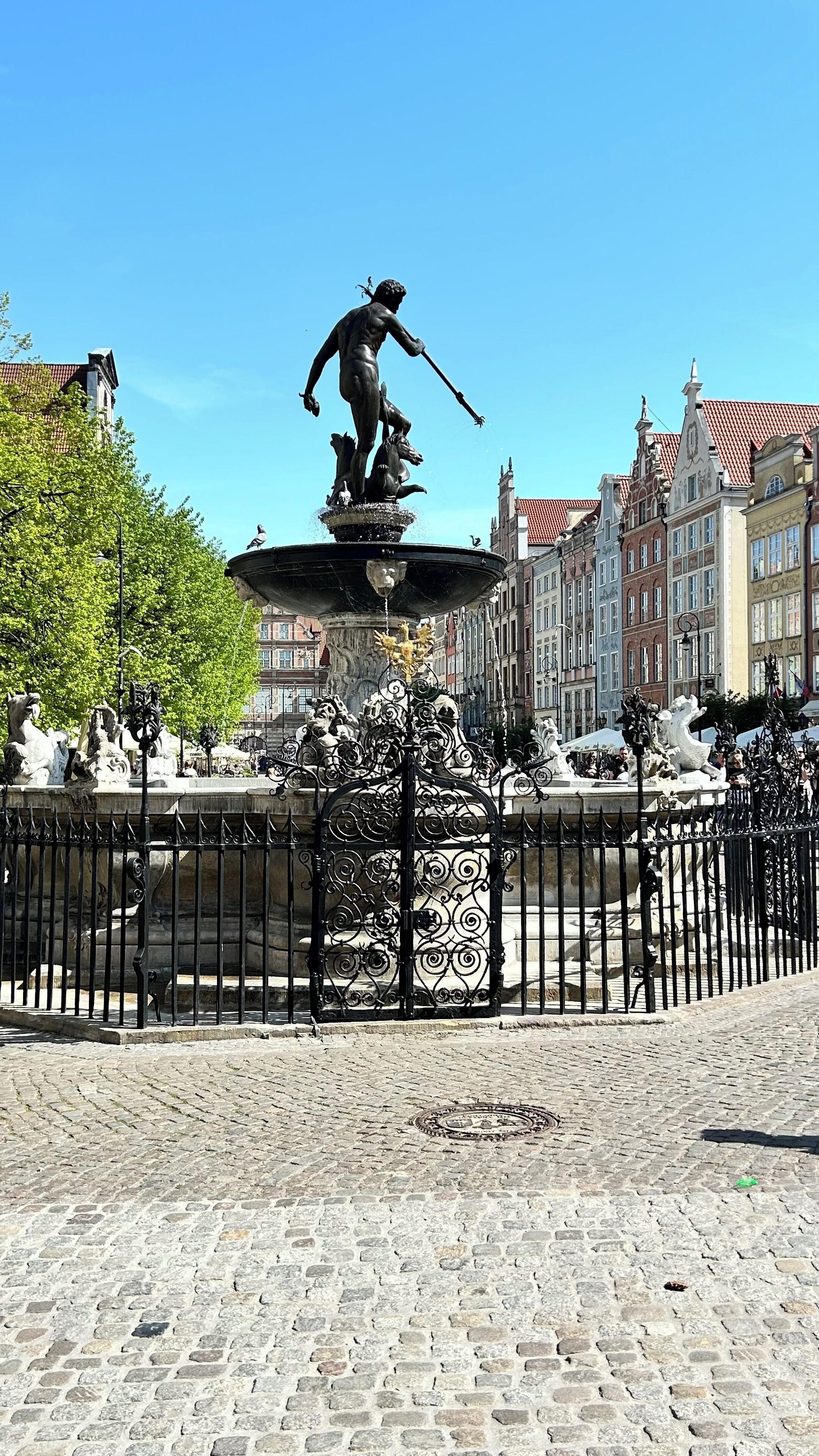 A historic fountain with statues, surrounded by cobblestone pavement and a wrought iron fence.