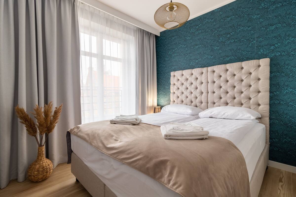A neatly made bed with a tufted headboard, a vase with dried plants, and a window with sheer curtains.