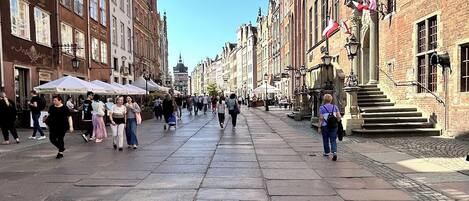 Une large rue piétonne bordée de bâtiments historiques, avec un espace ouvert central et des marches menant à un bâtiment surmonté d’une flèche.