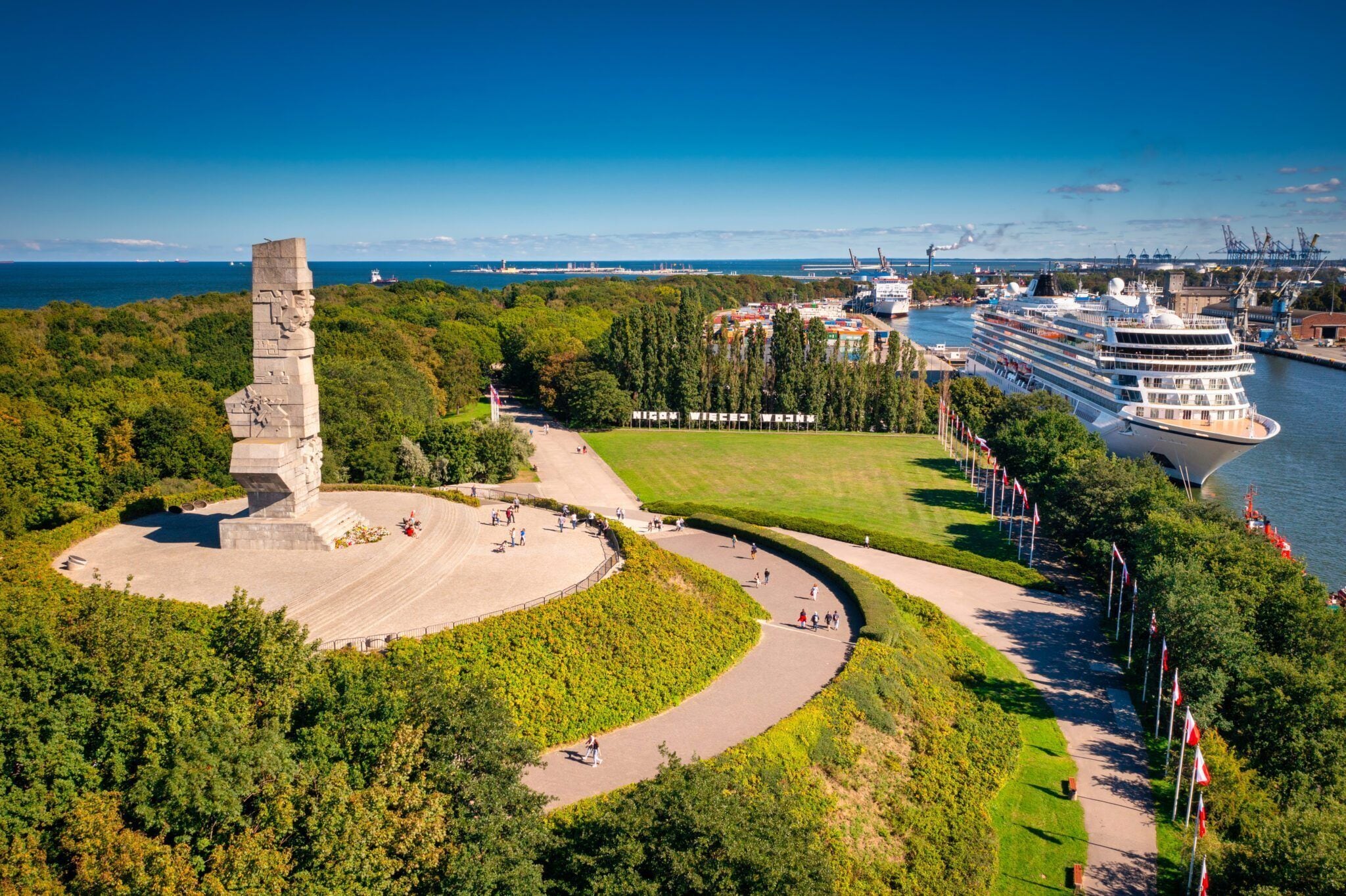 Zona costera con un monumento, un crucero amarrado y un parque arbolado.