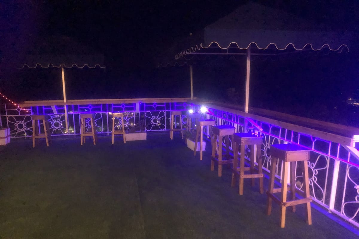An outdoor seating area with wooden stools and a railing, illuminated by purple lights.